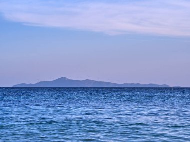 Mavi deniz ve gökyüzüne karşı rüzgar türbinleri olan uzak bir ada silueti, dağ çizgisi olan sakin bir deniz manzarası. Çevresel koruma.