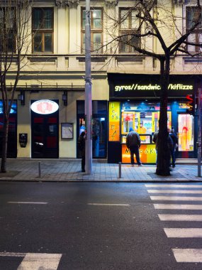 Belgrad, Sırbistan - 10 Ocak 2025: Sakin bir sokakta, bir jiroskop ve sandviç dükkanı da dahil olmak üzere aydınlanmış mağazaların kentsel gece manzarası