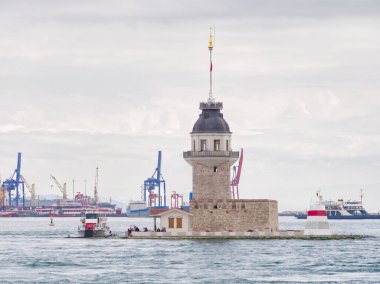 Arka planda gemi ve vinçlerin olduğu hareketli limanda antik kule deniz feneri