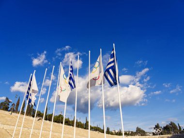 Panathenaic Stadyumu 'nda çok sayıda Yunan ve Olimpiyat bayrağı beyaz bulutlu, parlak mavi bir gökyüzüne karşı uzun bayrak direkleri sallıyor. Atina, Yunanistan