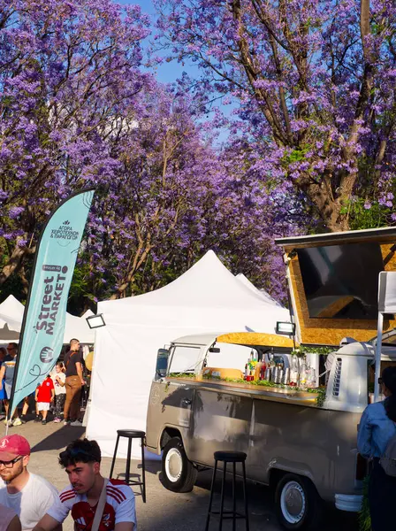 Atina, Yunanistan - 31 Mayıs 2025: Yemek kamyonu ve canlı mor jakaranda ağaçları altında çadırları olan açık hava pazarı