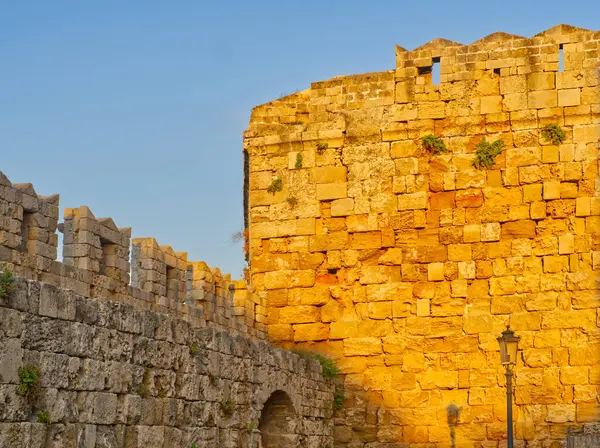 Altın güneş ışığı, Rodos 'taki ortaçağ taş tahkimatlarını aydınlatıyor. Çimenli duvarları ve çimenli hendek bölgesi olan eski bir kasaba.