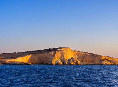 Derin mavi deniz sularından yükselen altın kireçtaşı uçurumlarının manzarası, sıcak günbatımı ışığıyla aydınlatılıyor. Koufonisia Adaları, Kiklad Adaları, Yunanistan