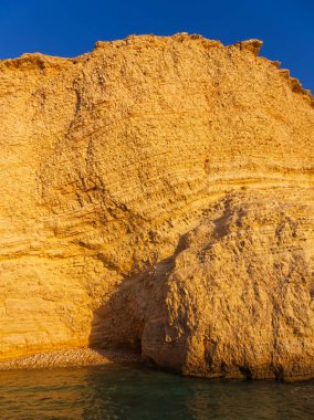 Temelinde mavi gökyüzü ve suya karşı kurulmuş doğal bir deniz mağarası olan görkemli altın kireçtaşı yüzeyi. Koufonisia Adaları, Kiklad Adaları, Yunanistan