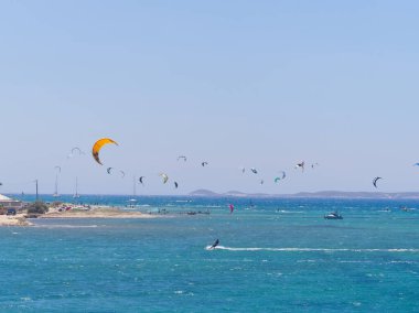 Çok renkli uçurtmalar gökyüzünü turkuaz bir denizin üstünde, uçurtmacılar rüzgarın ve dalgaların tadını çıkarıyorlar. Punta Limanı, Paros, Yunanistan