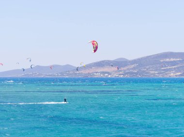 Uzak tepeleri ve gökyüzünde görülebilen büyük uçurtmaları olan canlı mavi okyanusta yalnız uçurtmacılar. Paros Adası, Yunanistan
