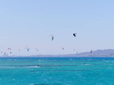 Çok renkli uçurtmalar gökyüzünü turkuaz bir denizin üstünde, uçurtmacılar rüzgarın ve dalgaların tadını çıkarıyorlar. Paros Adası, Yunanistan