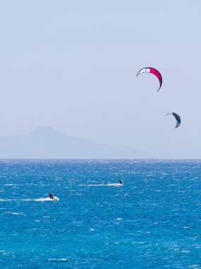 İki uçurtma sürücüsü canlı mavi bir deniz ve sisli ufukta renkli uçurtmalarla dalgalara biniyor. Punta Limanı, Paros, Yunanistan