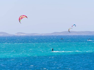 Uzak tepeleri ve gökyüzünde görülebilen büyük uçurtmaları olan canlı mavi okyanusta yalnız uçurtmacılar. Paros Adası, Yunanistan