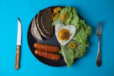 on a bright blue background, a black plate in which a hearty breakfast of scrambled eggs in the shape of a heart on healthy greens next to fragrant bread and vegetables and juicy sausages