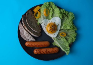 on a bright blue background, a black plate in which a hearty breakfast of scrambled eggs in the shape of a heart on healthy greens next to fragrant bread and vegetables and juicy sausages