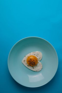 on a bright blue background, a black plate in which a hearty breakfast of scrambled eggs in the shape of a heart on healthy greens next to fragrant bread and vegetables and juicy sausages
