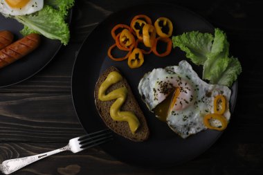 hearty healthy romantic breakfast of heart-shaped scrambled eggs and juicy sausages with green salad and fragrant bread in a black plate on a wooden surface