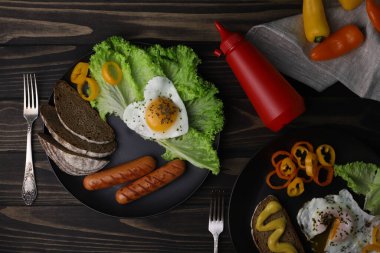 hearty healthy romantic breakfast of heart-shaped scrambled eggs and juicy sausages with green salad and fragrant bread in a black plate on a wooden surface