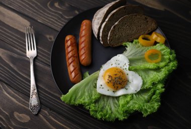 hearty healthy romantic breakfast of heart-shaped scrambled eggs and juicy sausages with green salad and fragrant bread in a black plate on a wooden surface
