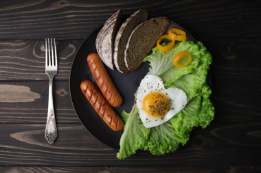 hearty healthy romantic breakfast of heart-shaped scrambled eggs and juicy sausages with green salad and fragrant bread in a black plate on a wooden surface