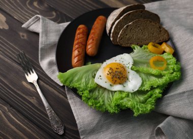 hearty healthy romantic breakfast of heart-shaped scrambled eggs and juicy sausages with green salad and fragrant bread in a black plate on a wooden surface