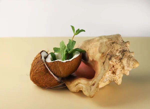 stock image two halves of a tropical coconut with white pulp and mint pieces in the middle next to seashells on a light background