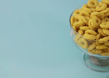 Kahvaltı için sağlıklı mısır gevrekleri. Narin bir zemin üzerinde şeffaf bir bardakta sarı halkalar şeklinde. Reklam etiketleri için ekranlara, afiş ilanlarına, reklamlara bakın.