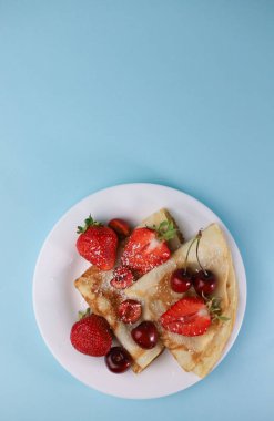 Lezzetli kokulu krepler, beyaz bir tabakta üçgen şeklinde katlanmış ve mavi arka planda lezzetli olgun çilekler, kırmızı kokulu kirazlar ve hindistan cevizi taneleriyle süslenmiş.