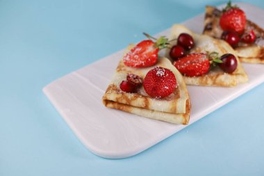 Lezzetli kokulu krepler, beyaz bir tabakta üçgen şeklinde katlanmış ve mavi arka planda lezzetli olgun çilekler, kırmızı kokulu kirazlar ve hindistan cevizi taneleriyle süslenmiş.