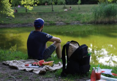 Yeşil suyla gölde yaz tatili pikniği. Piknikte, elinde olgun bir karpuz ve halının üstünde duran bir adam. Yanında siyah bir sırt çantası var.