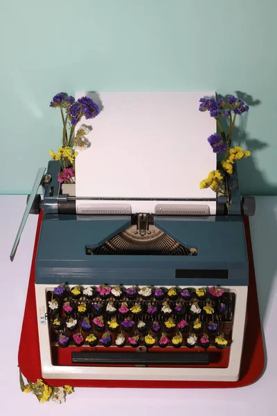 Retro style typewriter in dark gray and light gray with white sheet of paper. Bright flowers of different colors on the letters and on the typewriter. on a light blue background