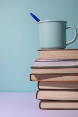 Mavi arka planda, üzerinde mavi bir kupa olan bir yığın kitap var.