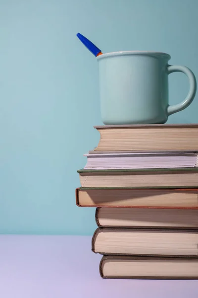 Mavi arka planda, üzerinde mavi bir kupa olan bir yığın kitap var.