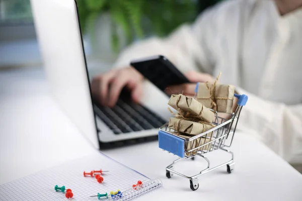 Minyatür mavi alışveriş arabalı online alışveriş konsepti beyaz dizüstü bilgisayarın önünde duruyor. İşçi elleri klavyede.