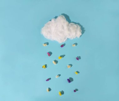 Mavi arka planda beyaz bir bulut var. Buluttan damlalar yerine çok renkli çiçekler düşüyor.