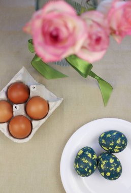 Paskalya yumurtaları beyaz bir tabağa boyanır. Sarı benekli yeşil yumurtalar. Yanında bir gül vazosu var.