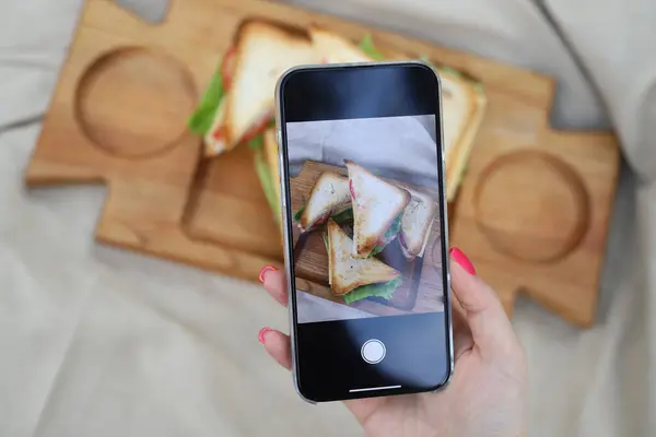 Blogcu telefonuyla yemek fotoğrafı çekiyor. Bir kadının eli telefonu tutar ve sandviçlerin fotoğrafını çeker.