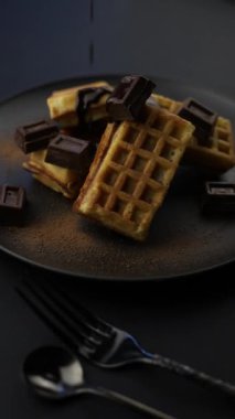 dikey video. Çikolatalı waffle 'lar siyah bir tabakta yatıyor. Çikolata parçaları ve sıcak çikolata bir dereye dökülüyor.