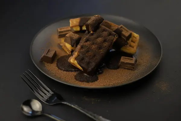 Dünya Çikolata Günü. Siyah bir tabakta kakao tozu serpiştirilmiş çikolata ve waffle parçaları..
