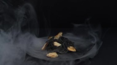 Siyah arka planda siyah tabakta midyeli siyah spagetti. Tabağın etrafında sigara iç