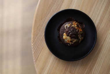 Tahta bir masanın üzerinde duran siyah bir tabağın üzerine fındık parçacıkları serpiştirilmiş çikolatalı kek.