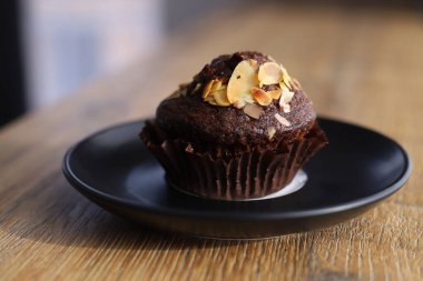 Tahta bir masanın üzerinde duran siyah bir tabağın üzerine fındık parçacıkları serpiştirilmiş çikolatalı kek.