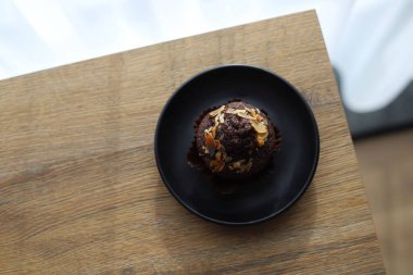 Tahta bir masanın üzerinde duran siyah bir tabağın üzerine fındık parçacıkları serpiştirilmiş çikolatalı kek.