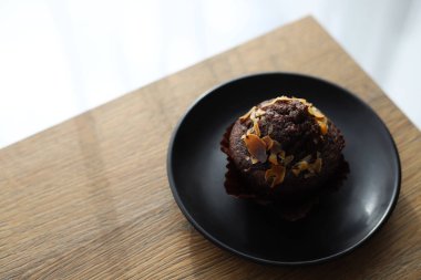 Tahta bir masanın üzerinde duran siyah bir tabağın üzerine fındık parçacıkları serpiştirilmiş çikolatalı kek.