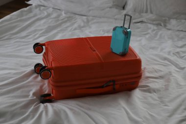 Turuncu büyük bir seyahat çantası ve beyaz çarşaflı bir yatağın arka planında küçük mavi bir bavul.