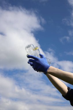 Çöp ve plastik. Doğadaki çimlerde çöp ve plastik şişeleri temizlemek. Mavi iş eldivenli birinin elleri mor bir çöp torbasında plastik şişe topluyor.