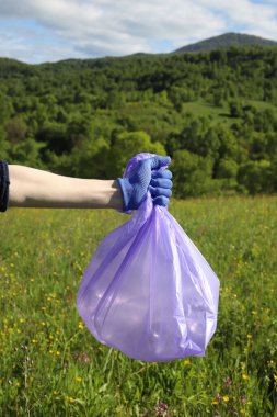 Çöp ve plastik. Doğadaki çimlerde çöp ve plastik şişeleri temizlemek. Mavi iş eldivenli birinin elleri mor bir çöp torbasında plastik şişe topluyor.