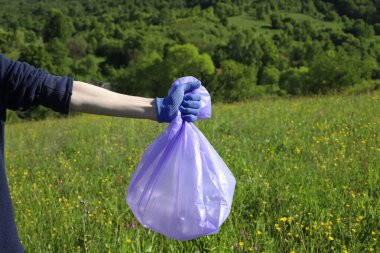 Çöp ve plastik. Doğadaki çimlerde çöp ve plastik şişeleri temizlemek. Mavi iş eldivenli birinin elleri mor bir çöp torbasında plastik şişe topluyor.