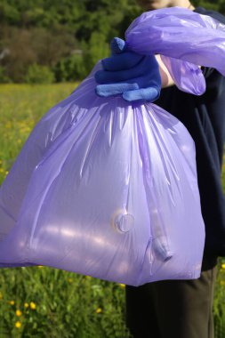 Çöp ve plastik. Doğadaki çimlerde çöp ve plastik şişeleri temizlemek. Mavi iş eldivenli birinin elleri mor bir çöp torbasında plastik şişe topluyor.