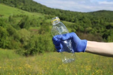 Çöp ve plastik. Doğadaki çimlerde çöp ve plastik şişeleri temizlemek. Mavi iş eldivenli birinin elleri mor bir çöp torbasında plastik şişe topluyor.