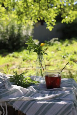 Doğal bir masada şeffaf bir fincanda çay. Yanında da kır çiçekleriyle şeffaf bir vazo var. Köyde yaz mevsimi