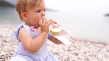 Küçük kız çakıl taşı plajında otururken biberondan limon suyu içiyor. Yüksek kalite 4k görüntü