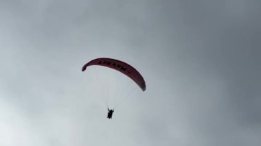 Bir insan gökyüzünde canlı kırmızı bir paraşütle yamaç paraşütü yapıyor. Rüzgâr sporlarının ve bulutların nefes kesen manzarasının tadını çıkarıyor.