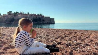 Genç bir kız kumlu sahilde oturuyor, mutlu bir şekilde elma yiyor ve suyun ufukla buluşmasını seyrediyor. Güneşli gökyüzü ve huzurlu manzara onun için eğlenceli ve kolay bir kaçış sağlar.